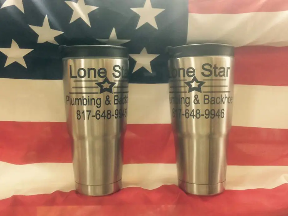 Two steel tumblers engraved Lone Star Plumbing & Backhoe with the phone number, resting on an American flag